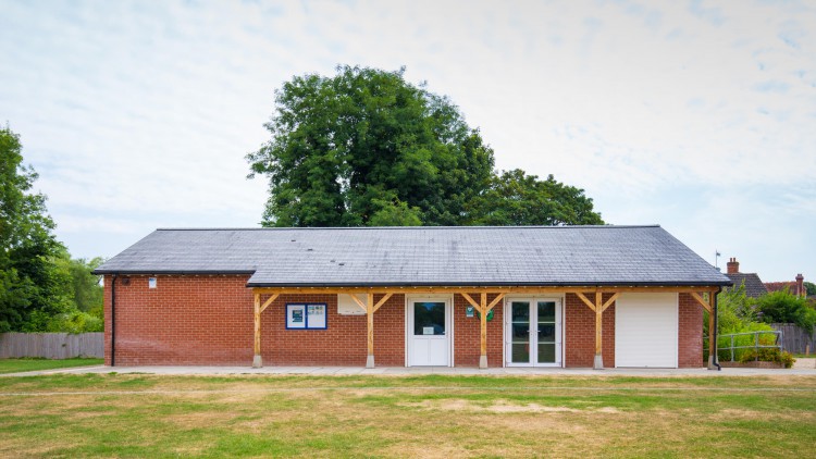 a straight shot of the pavilion which is a one level brick building with a grey roof and white doors and a small shelter in a big field 